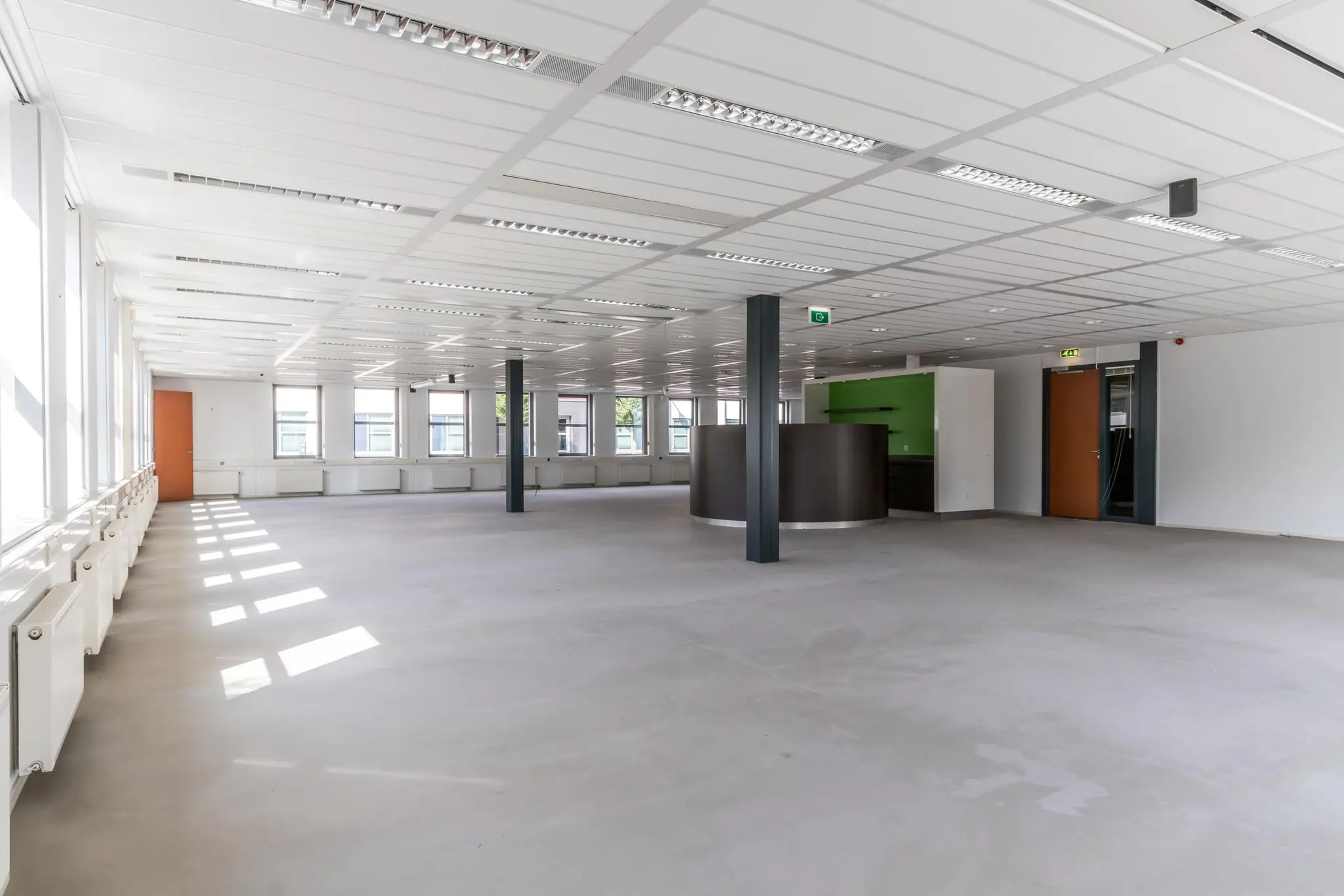 Lichte, lege kantoorruimte met grote ramen, systeemplafond en een ronde balie bij een groene wand.